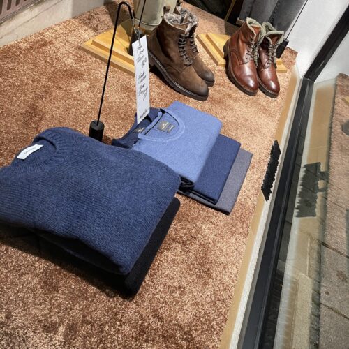 Zusammengelegte blaue Pullover, braune Lederschuhe und Preisschild auf braunem Teppich.